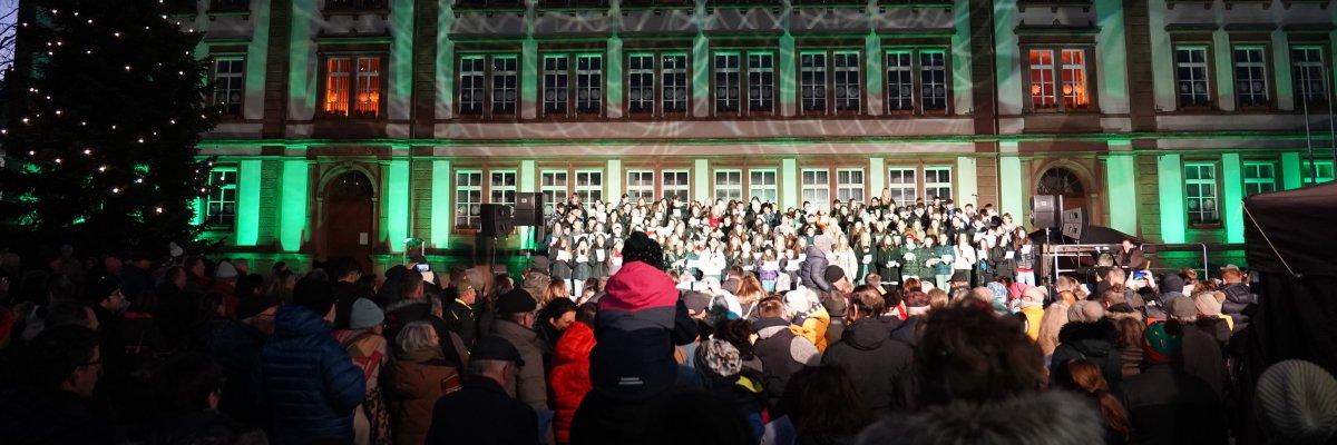 die Sänger stehen auf einer Bühne auf dem Exerzierplatz. Dahinter sieht man das Rathaus. 