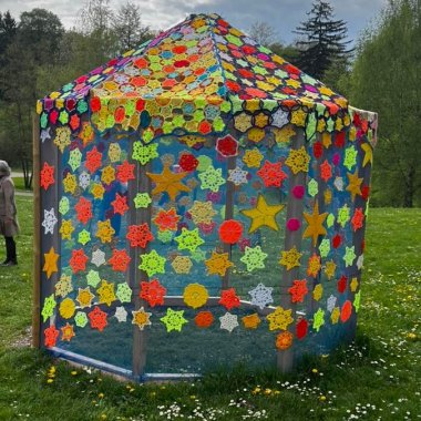 Ein sechseckiger Pavillon im Grünen, der über und über mit bunten gehäkelten Sternen versehen ist 