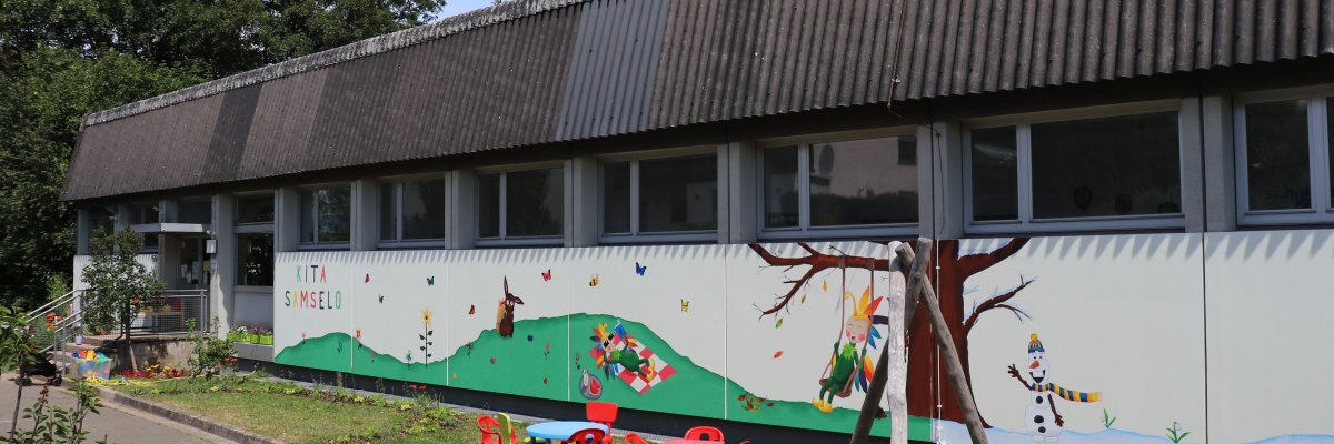 Abgebildet ist das Gebäude der Kita mit Wandgemälde (verschiedene Jahreszeitenmotive vom Samselo) 