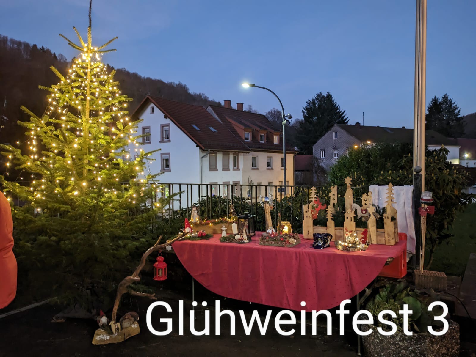 Ein Tisch mit roter Tischdecke, Darauf stehen verschiedene Dekoartikel aus Holz. Links neben dem Tisch befindet sich ein leuchtender Tannenbaum