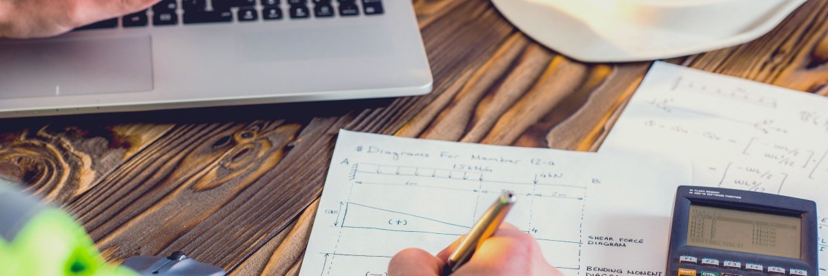 Mann zeichnet auf Block daneben ein Taschenrechner und im Hintergrund ein Laptop und Helm  