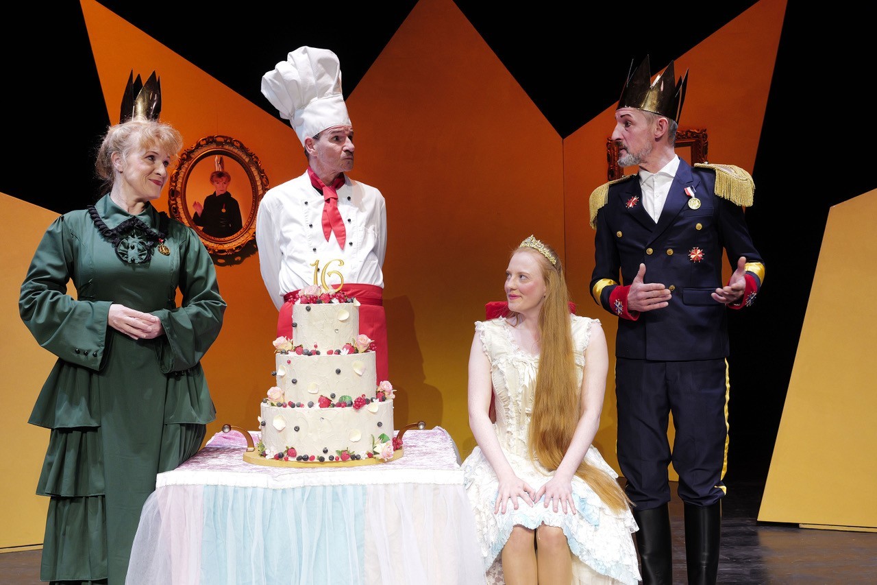4 Darsteller während einer Scene aus dem Stück Dornröschen. Die Prinzessin sitzt in der Mitte vor einem Kuchen während die Anderen 3 um sie herumstehen