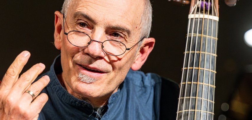 Ein älterer Mann mit kurzem grauem Haar und runder Brille sitzt mit einem historischen Streichinstrument, vermutlich einer Viola da gamba. Er trägt ein dunkelblaues Hemd, hält einen Bogen in der rechten Hand und gestikuliert mit der linken Hand, während er nach vorne blickt. Das Instrument steht aufrecht neben ihm und hat einen geschnitzten Kopf am oberen Ende des Halses. Im Hintergrund ist eine dunkle, unscharfe Bühne zu sehen.