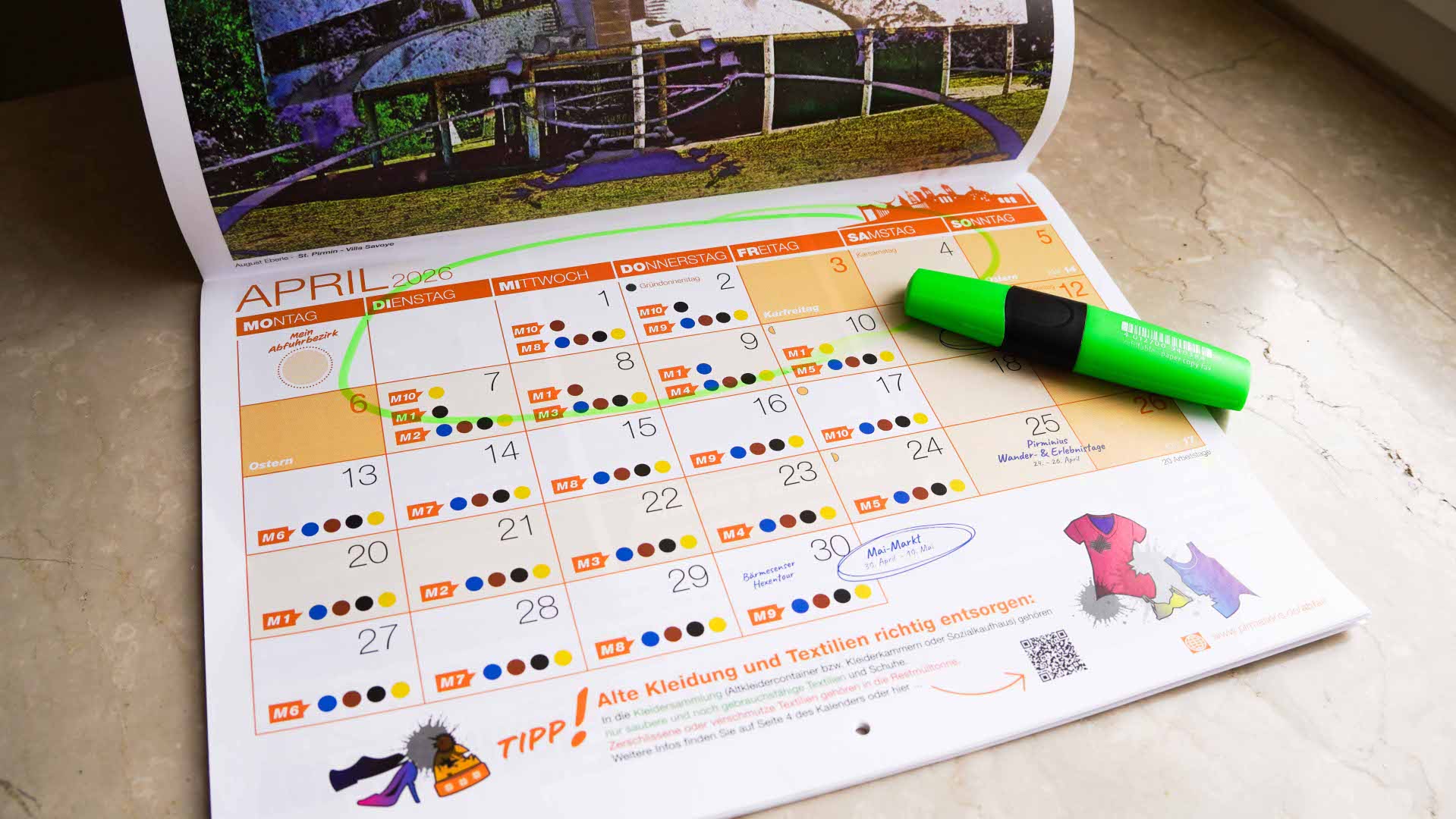 Abfallkalender Ein aufgeschlagener Kalender für den Monat April 2026 liegt auf einer Oberfläche. Mehrere Termine sind farbig markiert, und ein grüner Textmarker liegt quer auf der rechten Seite. Einzelne Daten sind eingekreist oder hervorgehoben.