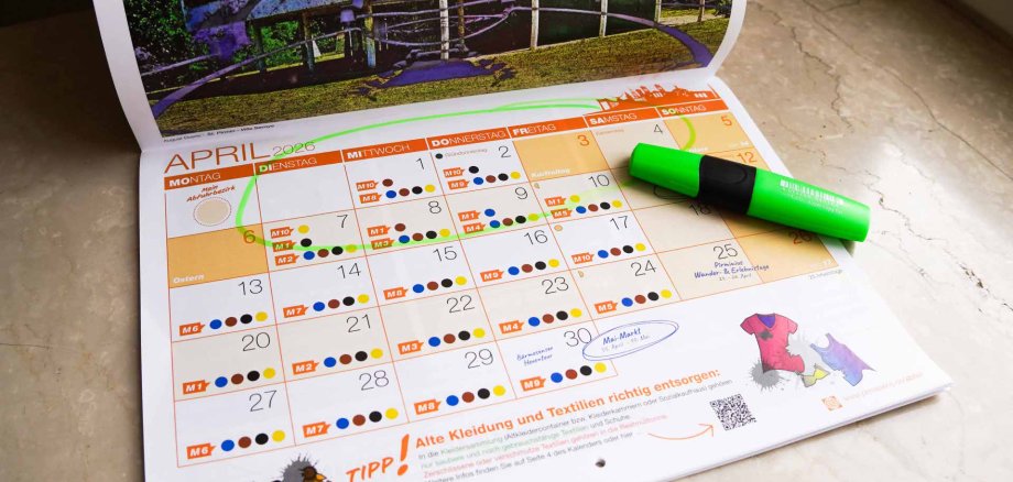 Abfallkalender Ein aufgeschlagener Kalender für den Monat April 2026 liegt auf einer Oberfläche. Mehrere Termine sind farbig markiert, und ein grüner Textmarker liegt quer auf der rechten Seite. Einzelne Daten sind eingekreist oder hervorgehoben.