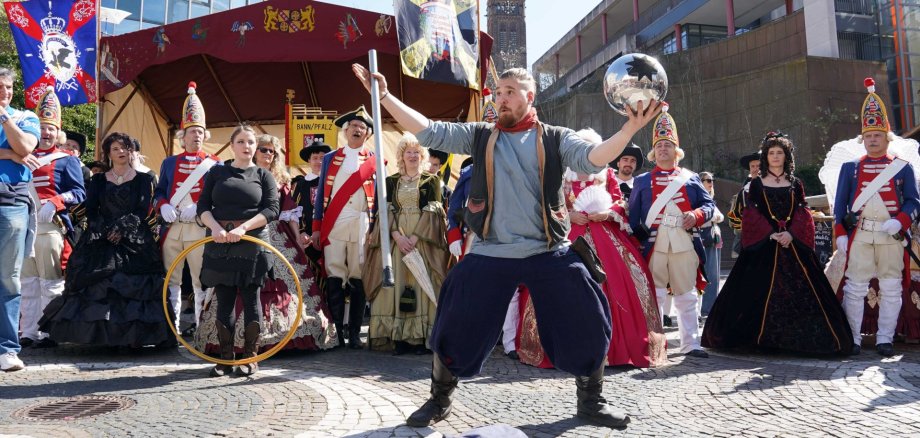 Auf dem Weg zum Schloßplatz: Landgraf Ludwig IX. und seine Gemahlin Caroline, begleitet von der CVP-Grenadiergarde. Ein Straßenkünstler in historischer Kleidung jongliert mit einer Kugel und einem Stab vor einer Gruppe von Menschen in aufwendigen Kostümen aus verschiedenen Epochen. Im Hintergrund stehen ein dekoriertes Zelt mit Wappen und Fahnen sowie moderne Gebäude; die Szene wirkt wie ein historisches Straßenfest oder eine Aufführung im Freien.
