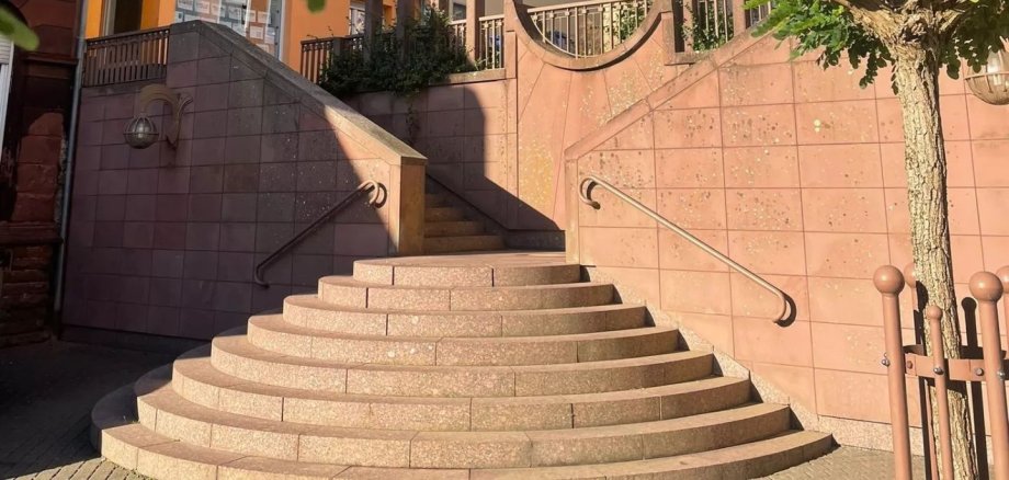 Kleine Treppenanlage mit halbkreisförmigen Stufen und Geländern führt zu einem erhöhten Platz vor Wohnhäusern; umgeben von Bäumen und blauem Himmel.