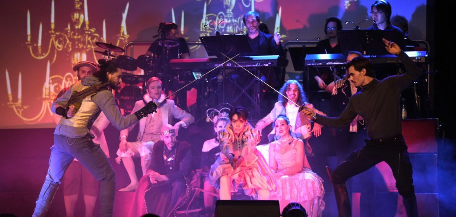 Theaterszene auf einer Bühne: Zwei Darsteller stehen sich mit Degen gegenüber und fechten, während eine Gruppe von Personen in historischen Kostümen im Hintergrund sitzt und zuschaut. Hinter ihnen spielt eine Band mit Instrumenten. Die Bühne ist farbig beleuchtet, und im Hintergrund sind große Projektionen von Kronleuchtern zu sehen.