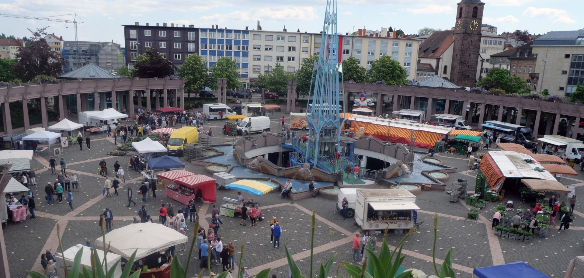 Blick von oben auf einen belebten Marktplatz mit vielen Ständen, Besuchern und einem Brunnen in der Mitte; im Vordergrund Blumen, im Hintergrund Gebäude und ein Kirchturm.