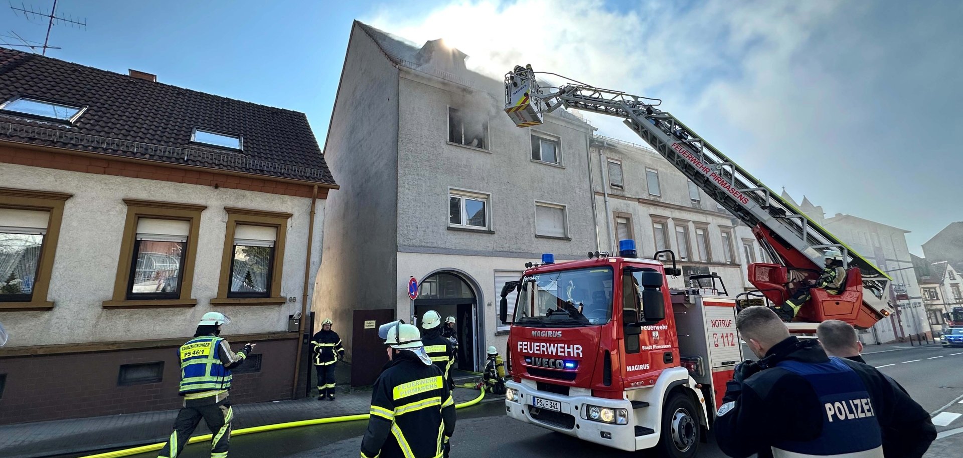Beim Eintreffen der Feuerwehr drang dichter schwarzer Rauch aus dem Fenster eines unbewohnten Mehrfamilienhauses in der Rodalber Straße. Die Feuerwehr brachte den Brand schnell unter Kontrolle und konnte ein Übergreifen der Flammen auf den Dachstuhl verhindern. Feuerwehreinsatz in einer Wohnstraße: Aus dem Dach eines mehrstöckigen Hauses dringt Rauch, während Einsatzkräfte mit Schläuchen und einer Drehleiter arbeiten. Ein Feuerwehrfahrzeug steht vor dem Gebäude, und ein Feuerwehrmann befindet sich im Korb der ausgefahrenen Leiter nahe dem Dach. Mehrere Feuerwehrleute und ein Polizist stehen auf der Straße und koordinieren den Einsatz.