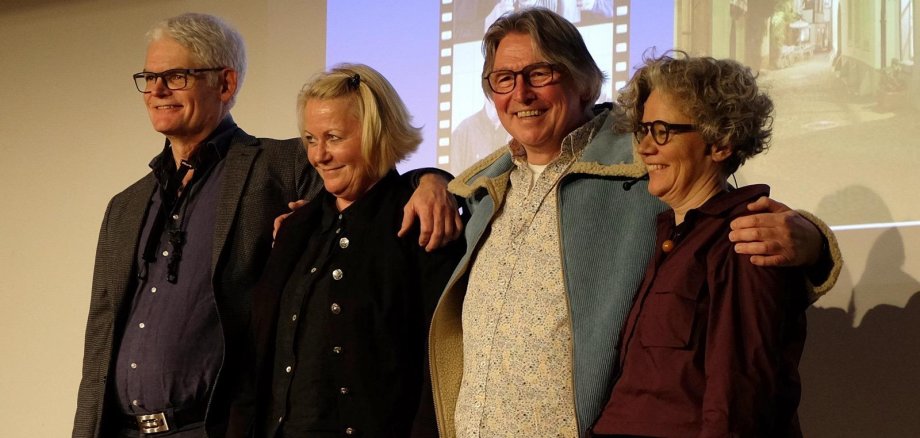 Vier Personen stehen nebeneinander auf einer Bühne, haben die Arme umeinander gelegt und lächeln in die Kamera. Sie tragen unterschiedliche Alltagskleidung, und im Hintergrund ist eine Projektionsfläche mit einem unscharfen Bild zu sehen.