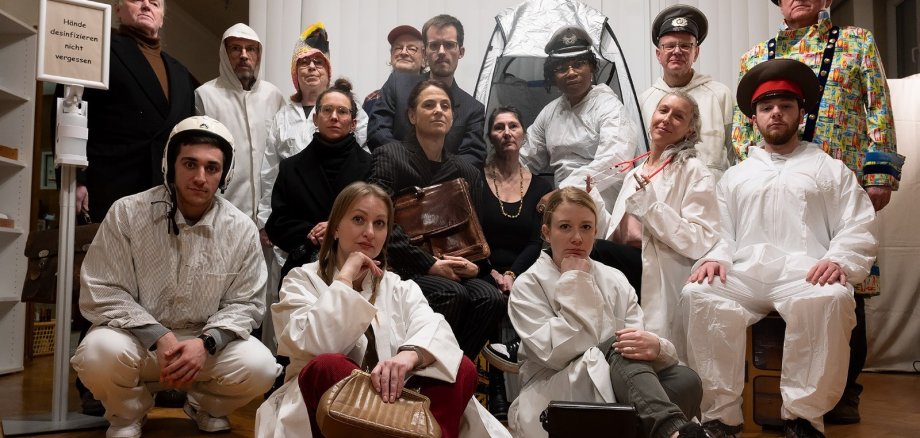 Gruppe von etwa 15 Personen in einem Innenraum, viele tragen weiße Schutzanzüge oder unterschiedliche Kostüme (z. B. Uniformen, Hut, bunte Kleidung). Einige sitzen vorne auf dem Boden oder auf Stühlen, andere stehen dahinter. Die Gruppe schaut ernst in die Kamera; links steht ein Desinfektionsspender mit dem Hinweis „Hände desinfizieren nicht vergessen“.