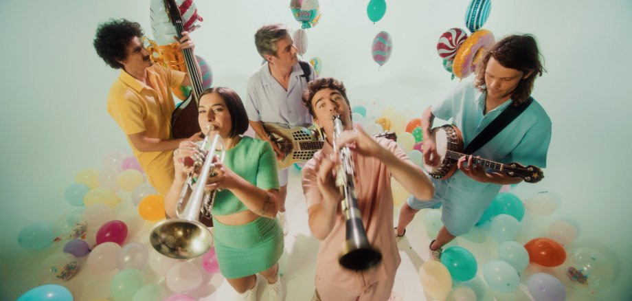Eine fröhliche Band spielt zwischen bunten Luftballons und Partydeko – zu sehen sind unter anderem Trompete, Klarinette, Kontrabass, Banjo und Resonatorgitarre in einer hellen Studio-Atmosphäre.