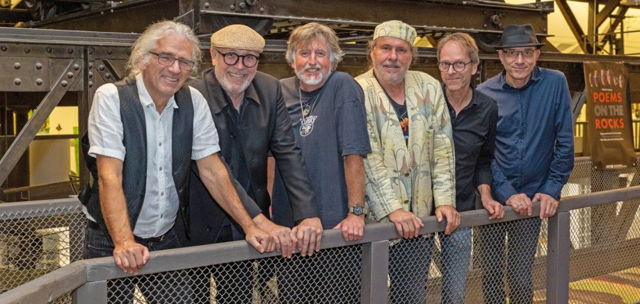 Sechs Mitglieder der Band „Poems on the Rocks“ stehen gemeinsam vor einer historischen Industrieanlage und lehnen sich lächelnd an ein Geländer. Im Hintergrund ist ein Banner der Band zu sehen, die Musiker tragen überwiegend dunkle Kleidung und verschiedene Hüte oder Mützen.