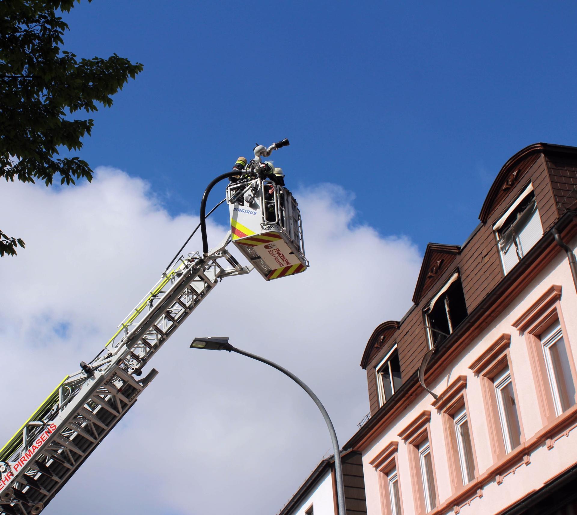 Feuerwehr-Drehleiter mit Korb und Wasserwerfer wird zu den oberen Stockwerken eines Gebäudes ausgefahren; mehrere Fenster sind geöffnet, Himmel im Hintergrund.