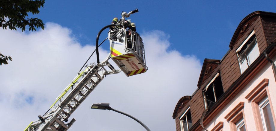 Feuerwehr-Drehleiter mit Korb und Wasserwerfer wird zu den oberen Stockwerken eines Gebäudes ausgefahren; mehrere Fenster sind geöffnet, Himmel im Hintergrund.