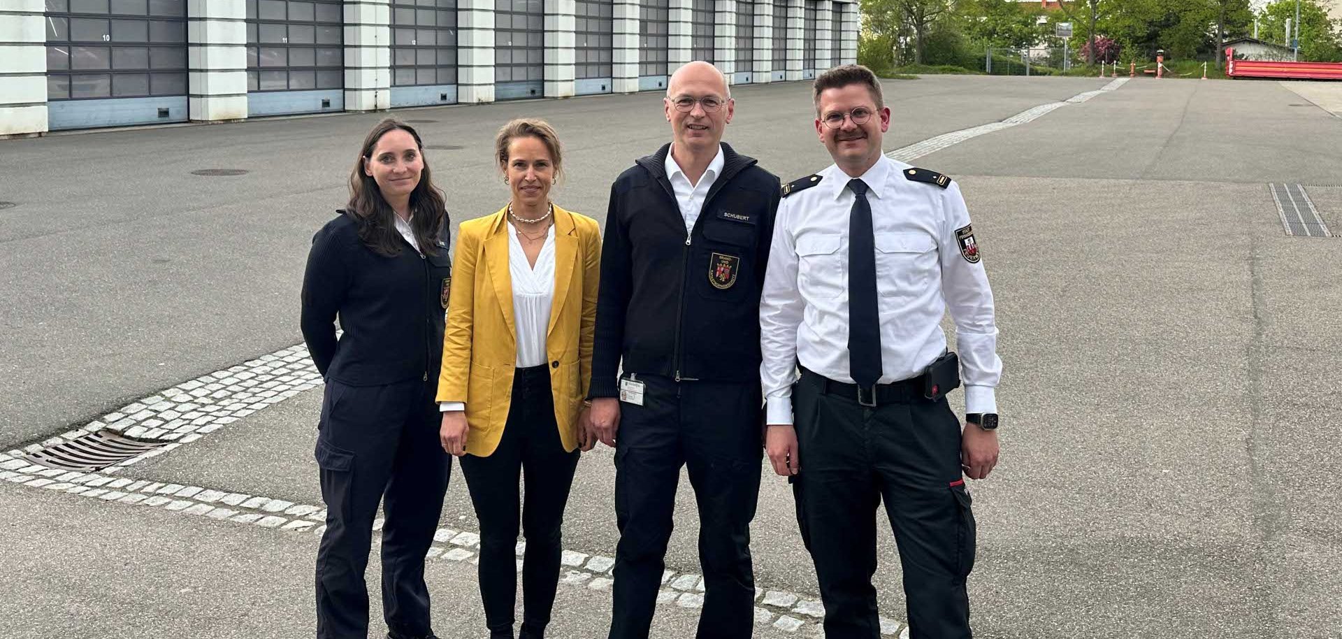 Vier Erwachsene stehen nebeneinander auf einem großen, offenen Gelände vor einer Reihe von Garagengebäuden; einige tragen Uniformen, im Hintergrund sind Bäume und ein bewölkter Himmel zu sehen.