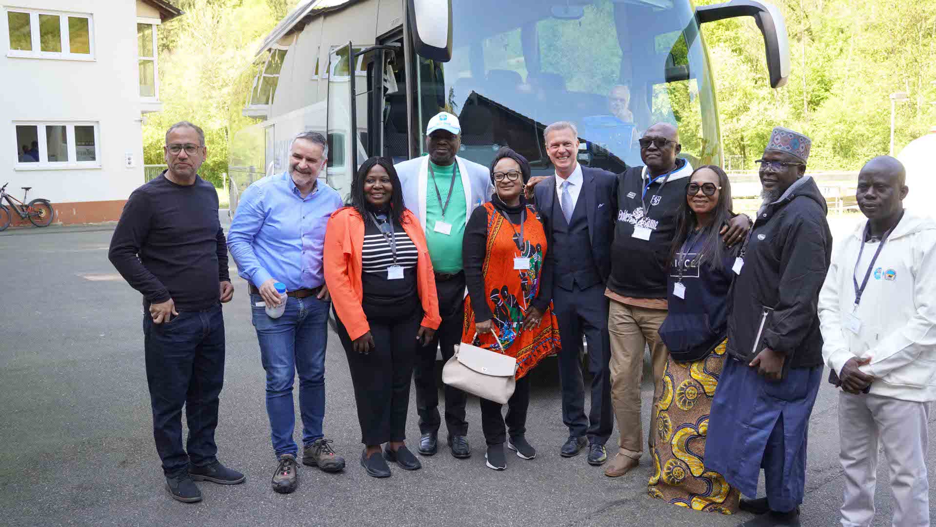Eine Gruppe von elf Erwachsenen steht lächelnd vor einem Reisebus im Freien; sie tragen unterschiedliche Kleidung, teils mit Namensschildern, und posieren gemeinsam für ein Gruppenfoto.