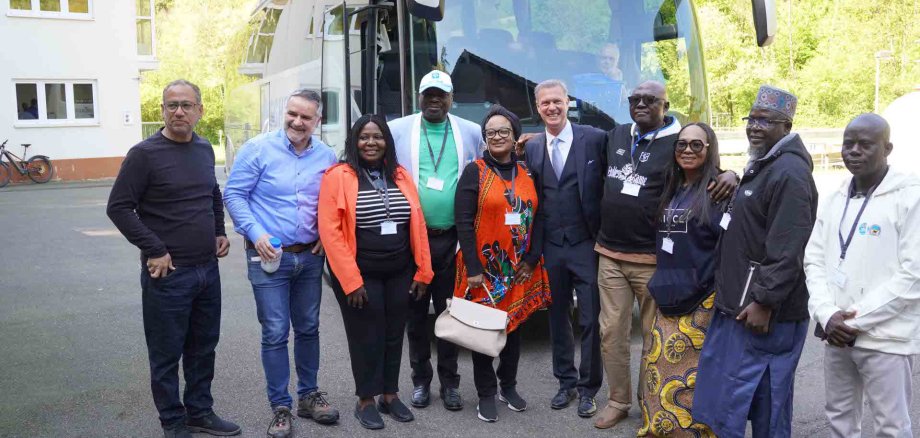 Eine Gruppe von elf Erwachsenen steht lächelnd vor einem Reisebus im Freien; sie tragen unterschiedliche Kleidung, teils mit Namensschildern, und posieren gemeinsam für ein Gruppenfoto.