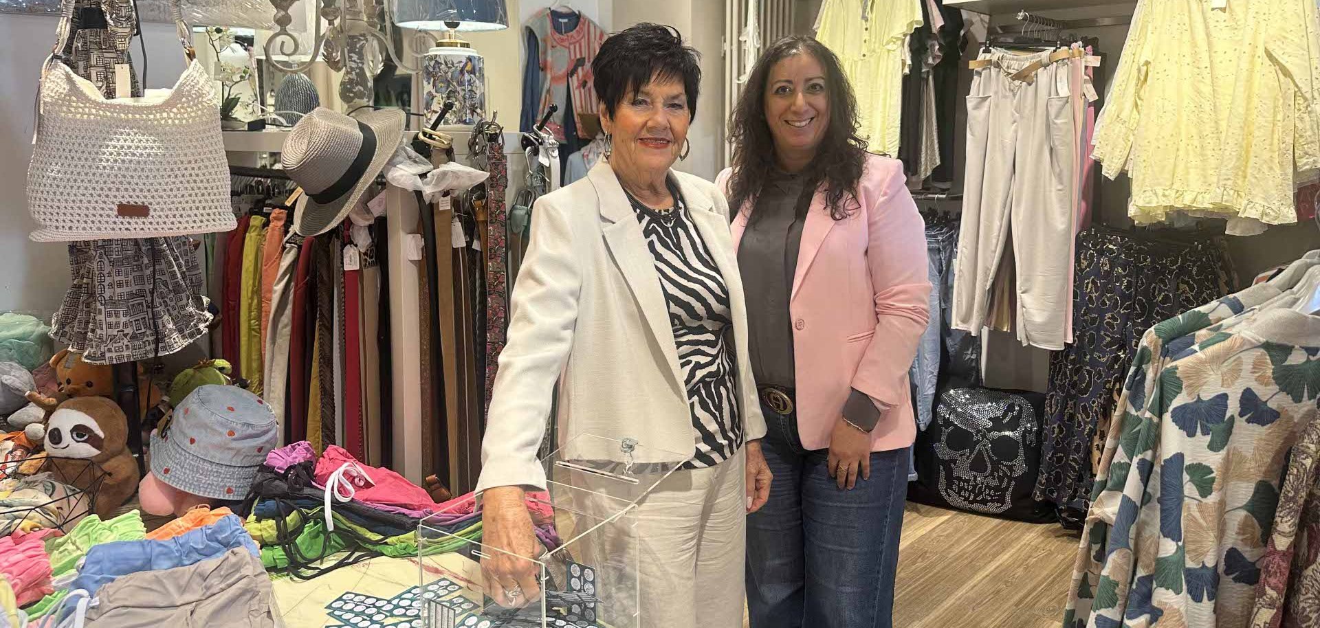Zwei Frauen stehen lächelnd in einem kleinen Bekleidungsgeschäft zwischen Kleiderständern und Accessoires; auf einem Tisch vor ihnen steht eine transparente Box mit Karten oder Losen.
