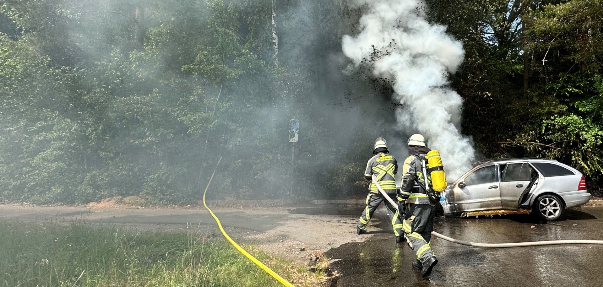 Die Feuerwehr Pirmasens hat am Muttertag einen Pkw-Brand auf der B10 / Abfahrt Haseneck erfolgreich bekämpft. Zwei Feuerwehrleute löschen ein brennendes Auto am Waldrand; dichter Rauch steigt über dem Fahrzeug auf.