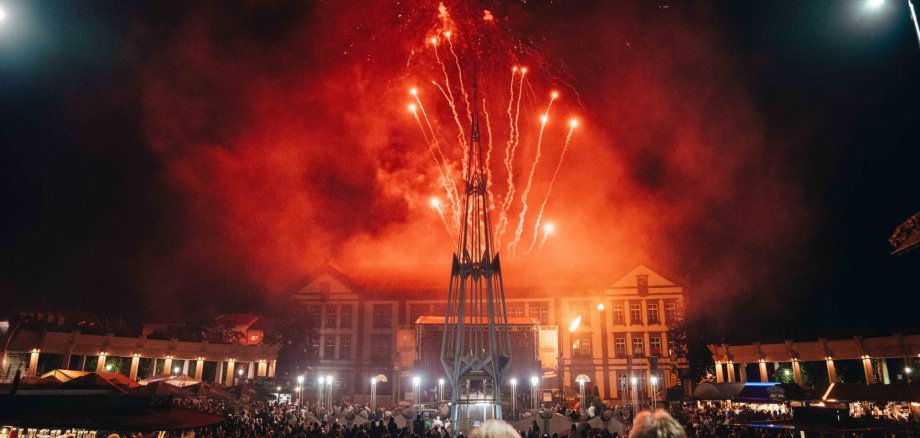 Feuerwerk Exerzierplatz Auf dem Foto ist ein großes Feuerwerk über einem gut besuchten Stadtplatz bei Nacht zu sehen. Leuchtend rote und orangefarbene Feuerwerkskörper steigen hinter einer markanten Skulptur in den Himmel und tauchen die Szenerie in ein warmes Licht. Vor einer historischen Gebäudekulisse haben sich zahlreiche Besucherinnen und Besucher versammelt, um das Spektakel zu verfolgen. Die Aufnahme vermittelt eine festliche Atmosphäre und fängt den Höhepunkt einer öffentlichen Veranstaltung eindrucksvoll ein.