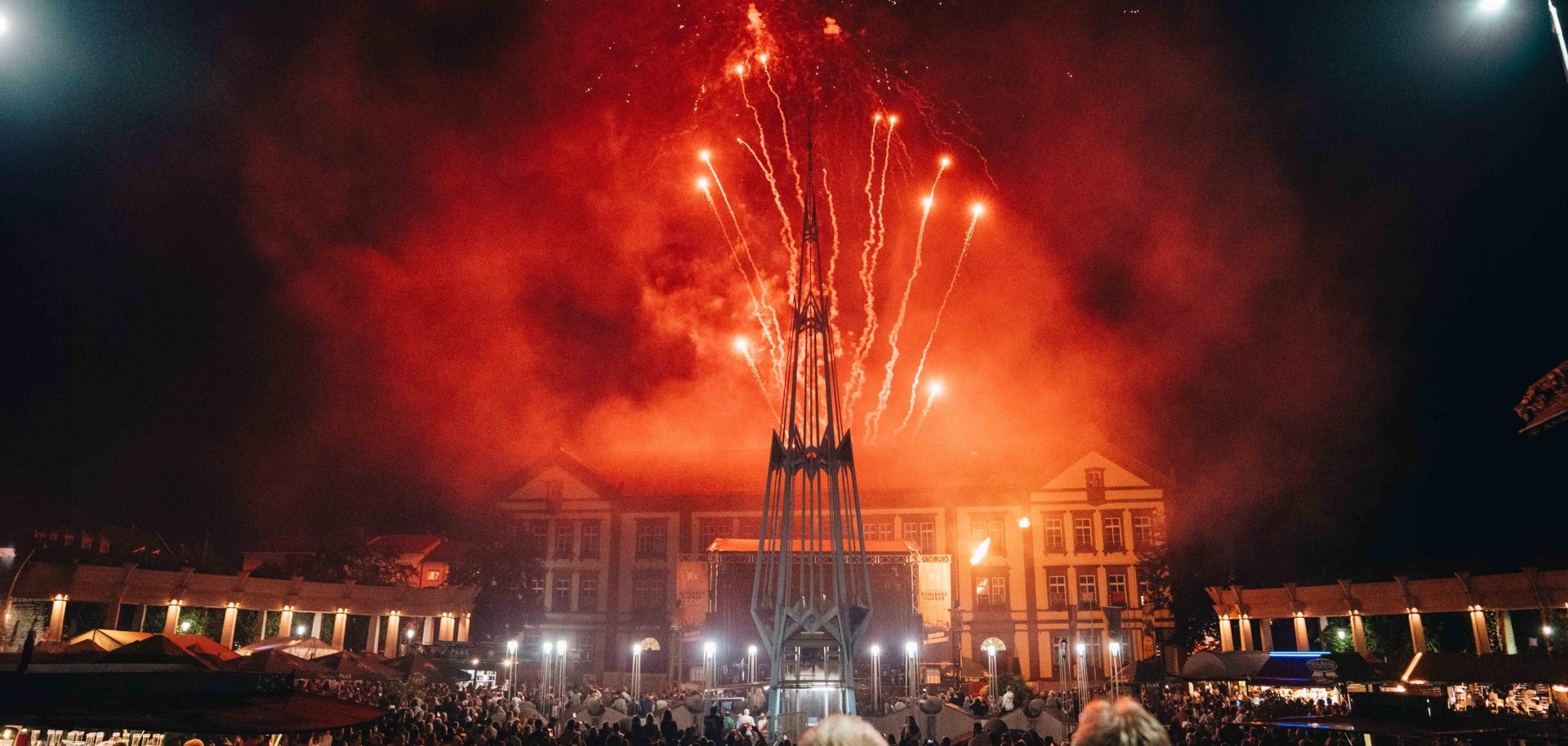 Feuerwerk Exerzierplatz Auf dem Foto ist ein großes Feuerwerk über einem gut besuchten Stadtplatz bei Nacht zu sehen. Leuchtend rote und orangefarbene Feuerwerkskörper steigen hinter einer markanten Skulptur in den Himmel und tauchen die Szenerie in ein warmes Licht. Vor einer historischen Gebäudekulisse haben sich zahlreiche Besucherinnen und Besucher versammelt, um das Spektakel zu verfolgen. Die Aufnahme vermittelt eine festliche Atmosphäre und fängt den Höhepunkt einer öffentlichen Veranstaltung eindrucksvoll ein.