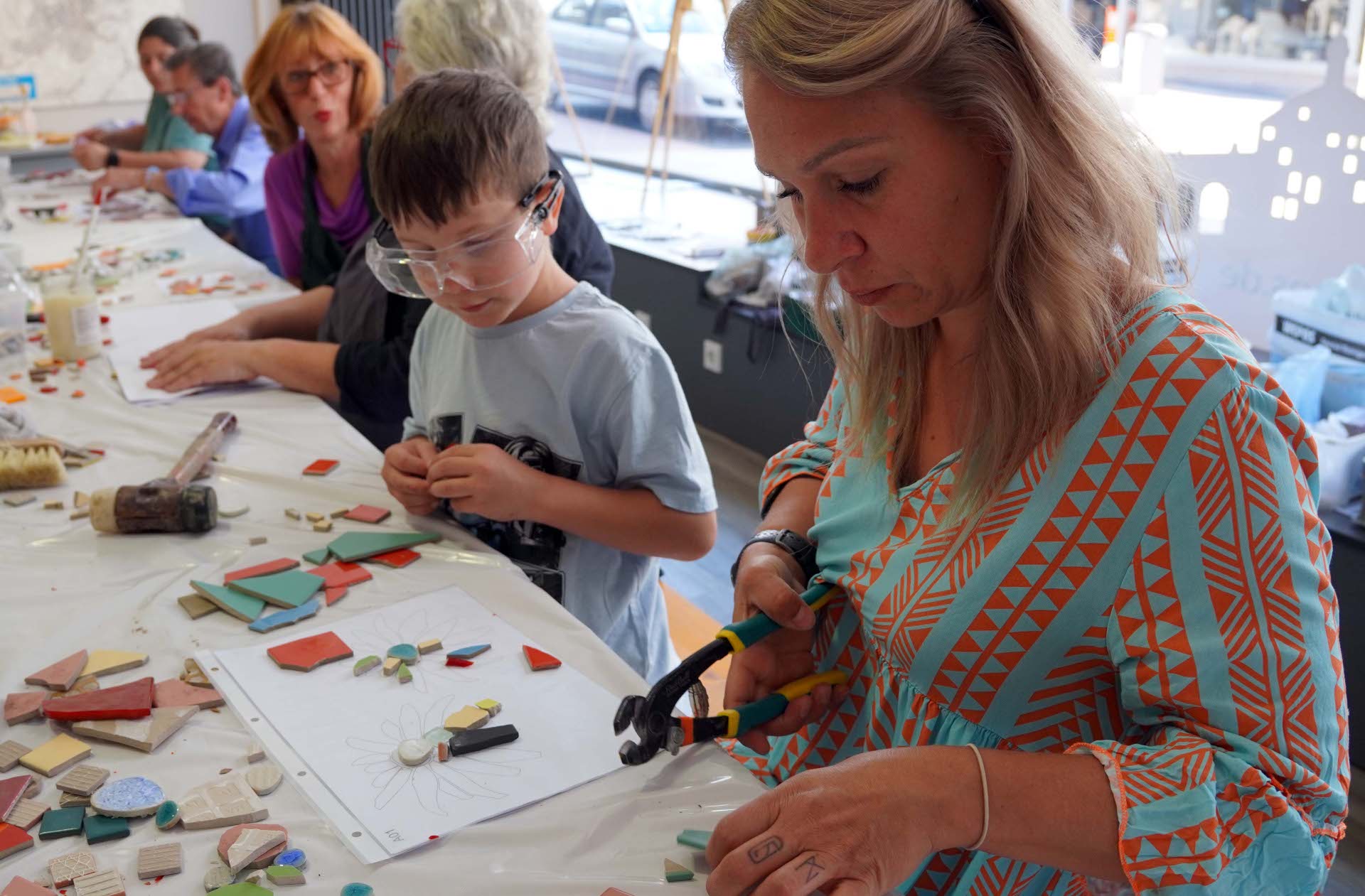 Das Bild zeigt mehrere Menschen, die gemeinsam an einem Mosaik-Workshop teilnehmen. Im Vordergrund schneidet eine Frau mit einer Mosaikzange bunte Keramikstücke zurecht. Neben ihr arbeitet ein Junge mit Schutzbrille konzentriert an einem Blumenmotiv, das auf einem Blatt vorgezeichnet ist. Auf dem Tisch liegen zahlreiche farbige Mosaiksteine, Werkzeuge und weitere Materialien. Im Hintergrund sitzen weitere Teilnehmerinnen und Teilnehmer an einer langen Tafel und gestalten ihre eigenen Werke. Die helle, kreative Atmosphäre vermittelt den Eindruck eines generationsübergreifenden Mitmachangebots.