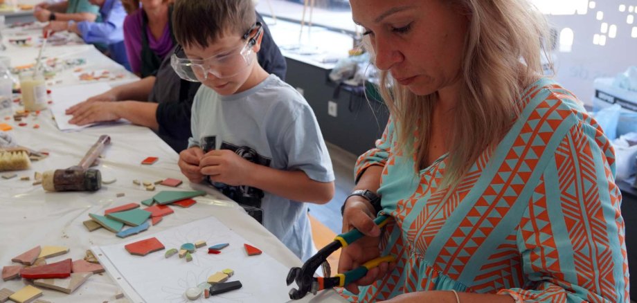 Das Bild zeigt mehrere Menschen, die gemeinsam an einem Mosaik-Workshop teilnehmen. Im Vordergrund schneidet eine Frau mit einer Mosaikzange bunte Keramikstücke zurecht. Neben ihr arbeitet ein Junge mit Schutzbrille konzentriert an einem Blumenmotiv, das auf einem Blatt vorgezeichnet ist. Auf dem Tisch liegen zahlreiche farbige Mosaiksteine, Werkzeuge und weitere Materialien. Im Hintergrund sitzen weitere Teilnehmerinnen und Teilnehmer an einer langen Tafel und gestalten ihre eigenen Werke. Die helle, kreative Atmosphäre vermittelt den Eindruck eines generationsübergreifenden Mitmachangebots.