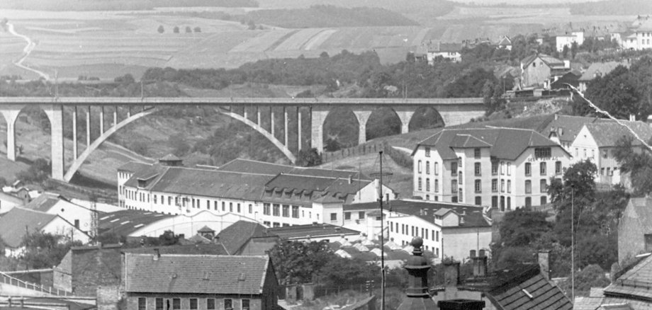 Das Bild zeigt eine historische Schwarz-Weiß-Aufnahme eines Stadtteils mit einer markanten Talbrücke im Mittelpunkt. Die mehrbogige Brücke überspannt ein weites Tal und verbindet zwei Anhöhen miteinander. Im Vordergrund sind zahlreiche Gebäude zu sehen, darunter größere Fabrik- oder Gewerbebauten sowie Wohnhäuser. Im Hintergrund erstrecken sich Felder, Wiesen und sanfte Hügel, die die ländliche Umgebung der Stadt erkennen lassen. Das Foto weist altersbedingte Gebrauchsspuren wie Kratzer und Flecken auf und vermittelt einen Eindruck der Stadt- und Verkehrsinfrastruktur vergangener Jahrzehnte.
