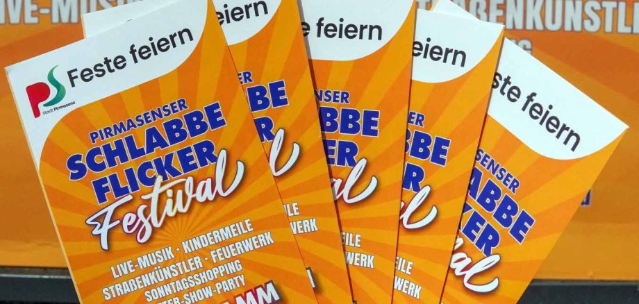 Kompakt und übersichtlich: Das Programmheft zum Schlabbeflicker-Festival 2026 ist ab sofort erhältlich. Das Bild zeigt eine Hand, die mehrere Programmflyer für das „Pirmasenser Schlabbe Flicker Festival“ auffächert. Im Hintergrund ist ein großes Plakat mit dem Festival-Logo in Orange und Gelb zu sehen. Auf den Flyern sind die Programmpunkte „Live-Musik“, „Kindermile“, „Straßenkünstler“, „Feuerwerk“, „Sonntagsshopping“ und „After-Show-Party“ sowie der Veranstaltungszeitraum vom 31. Juli bis 2. August 2026 angegeben. Die Aufnahme macht auf das Veranstaltungsprogramm aufmerksam und vermittelt Vorfreude auf das Stadtfest.
