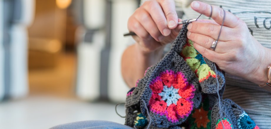 Nahaufnahme von Händen, die mit einer Häkelnadel an einem bunten Granny-Square-Projekt arbeiten. Die Person sitzt entspannt, während sie mit grauer Wolle ein farbenfrohes Häkelstück weiterfertigt.