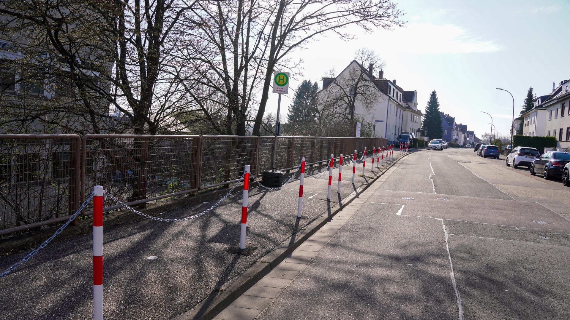 Eine Straße in einem Wohngebiet mit parkenden Autos auf der rechten Seite. Links verläuft ein Gehweg entlang eines Geländers, der durch rot-weiße Poller und eine Kette vom Fahrbahnrand getrennt ist. In der Mitte steht ein Haltestellenschild, das auf eine Bushaltestelle hinweist. Bäume und Wohnhäuser säumen die Straße.