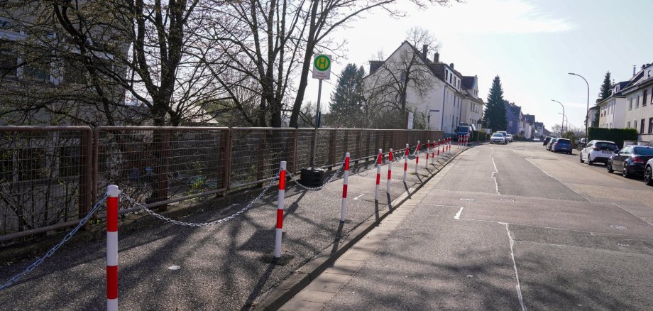 Eine Straße in einem Wohngebiet mit parkenden Autos auf der rechten Seite. Links verläuft ein Gehweg entlang eines Geländers, der durch rot-weiße Poller und eine Kette vom Fahrbahnrand getrennt ist. In der Mitte steht ein Haltestellenschild, das auf eine Bushaltestelle hinweist. Bäume und Wohnhäuser säumen die Straße.