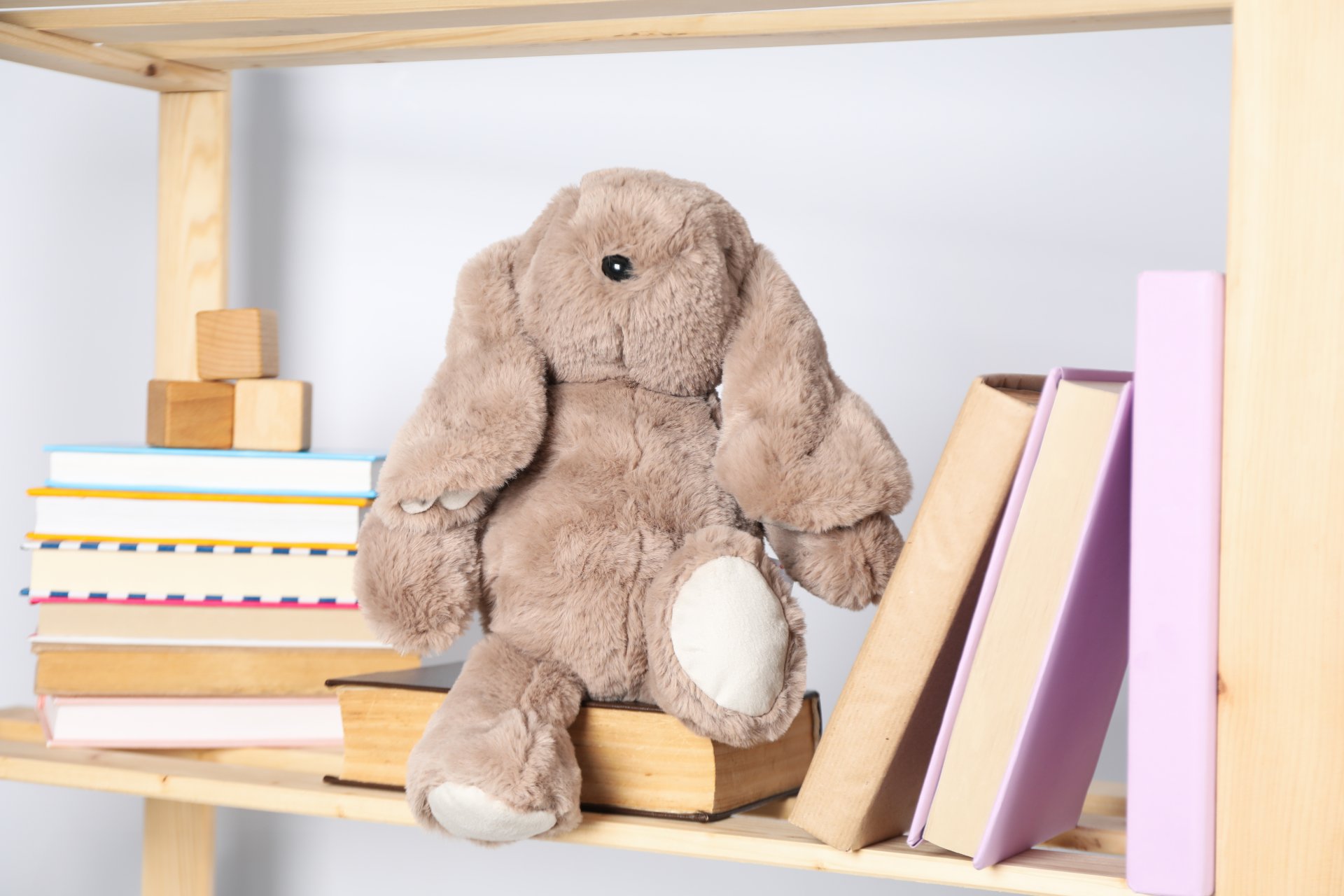 Ein braunes Plüschtier in Form eines Hasen sitzt auf einem Holzregal zwischen mehreren Büchern. Links liegen gestapelte Bücher und kleine Holzklötze, rechts stehen Bücher aufrecht. Der Hintergrund ist hell und neutral gehalten.