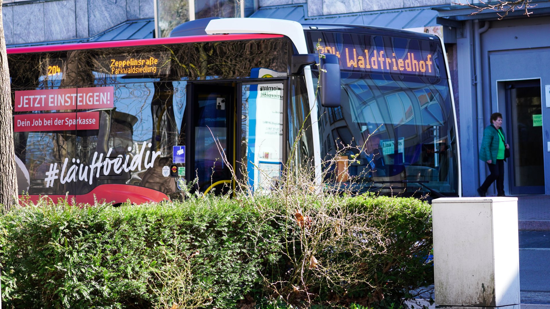 Das Bild zeigt einen modernen Linienbus des Stadtverkehrs Pirmasens an einer Haltestelle im Stadtgebiet. Auf der digitalen Zielanzeige sind die Fahrtziele „Zeppelinstraße / Parkwaldsiedlung“ sowie „Waldfriedhof“ zu erkennen. An der Fahrzeugseite befindet sich eine Werbefläche der Sparkasse mit dem Slogan „Jetzt einsteigen! Dein Job bei der Sparkasse“ und dem Hashtag „#läuftbeidir“. Im Vordergrund begrenzt eine Hecke den Blick auf den Bus, während rechts eine Passantin ein Gebäude betritt. Die Aufnahme vermittelt den öffentlichen Nahverkehr im Stadtzentrum.