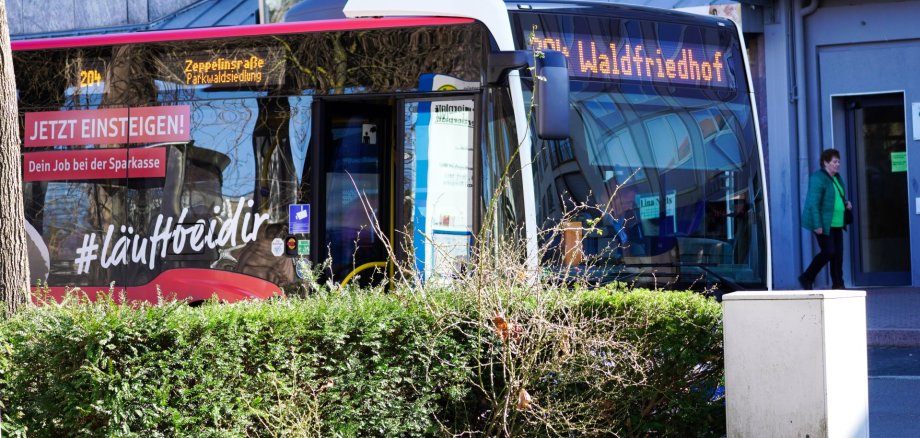 Das Bild zeigt einen modernen Linienbus des Stadtverkehrs Pirmasens an einer Haltestelle im Stadtgebiet. Auf der digitalen Zielanzeige sind die Fahrtziele „Zeppelinstraße / Parkwaldsiedlung“ sowie „Waldfriedhof“ zu erkennen. An der Fahrzeugseite befindet sich eine Werbefläche der Sparkasse mit dem Slogan „Jetzt einsteigen! Dein Job bei der Sparkasse“ und dem Hashtag „#läuftbeidir“. Im Vordergrund begrenzt eine Hecke den Blick auf den Bus, während rechts eine Passantin ein Gebäude betritt. Die Aufnahme vermittelt den öffentlichen Nahverkehr im Stadtzentrum.