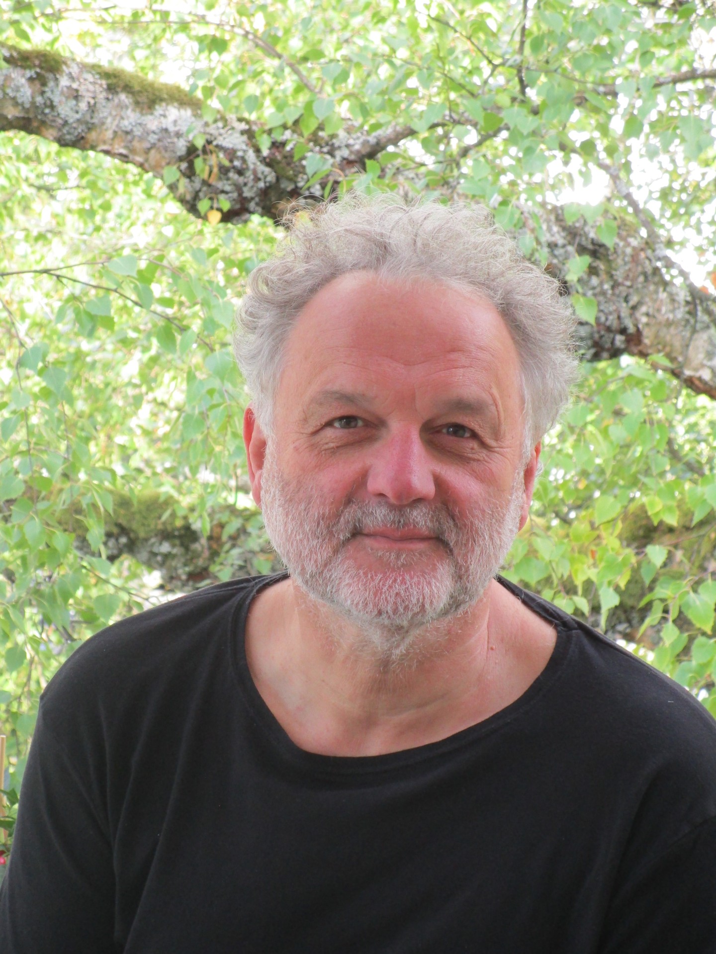 Dr. Rainer Metzger hält am 20. Mai 2026 den Vortrag „Bilderwelten der Weimarer Republik“ Porträt eines Mannes mit grauem lockigem Haar und Bart in schwarzem Shirt; im Hintergrund sind grüne Blätter und Äste eines Baumes zu sehen.
