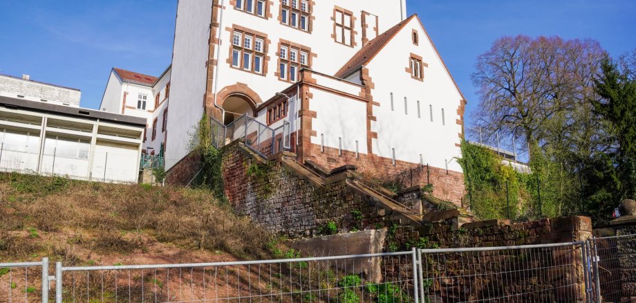 Ein großes, hell verputztes Gebäude mit roten Sandsteinelementen steht auf einem Hang. Eine steinerne Treppe führt von unten zu einem erhöhten Eingang mit Rundbogen. Vor dem Hang verläuft ein Metallzaun, und im Hintergrund sind Bäume sowie ein klarer blauer Himmel zu sehen.