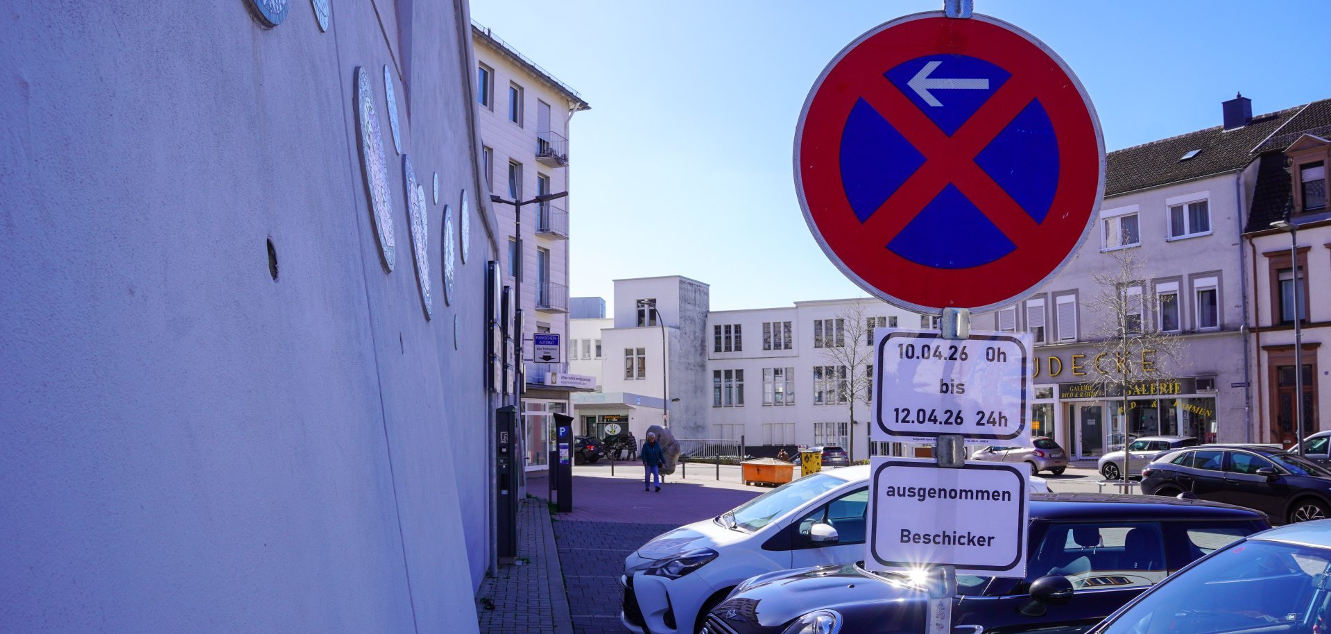 Parkverbotsschild mit Zusatzhinweis auf eine zeitlich begrenzte Regelung (10.04. bis 12.04.) steht an einem Parkplatz mit mehreren Autos. Im Hintergrund sind Gebäude und eine kleine Platzsituation in einer Stadt zu sehen.