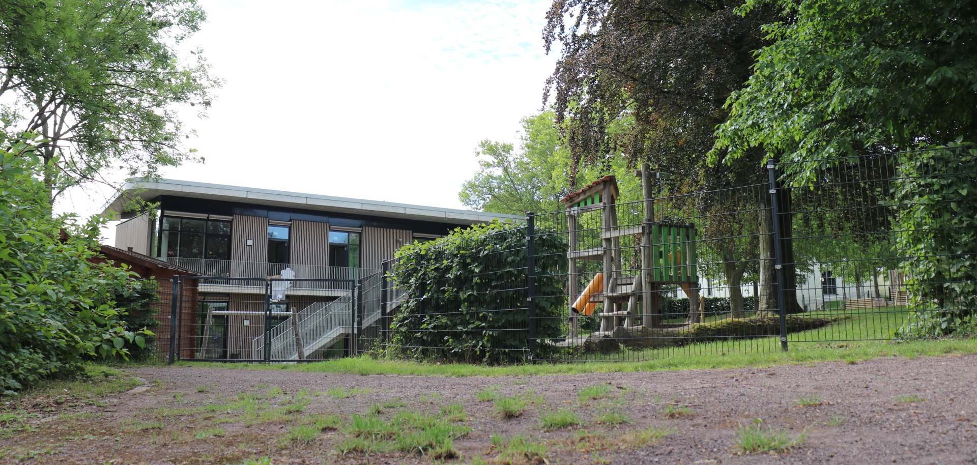 Ein modernes, zweigeschossiges Gebäude mit großen Fenstern steht hinter einem umzäunten Außengelände. Rechts befindet sich ein Spielplatz mit Klettergerüst und Rutsche, umgeben von Bäumen und Grünflächen. Im Vordergrund ist ein unbefestigter Weg mit Gras zu sehen, während dichte Vegetation und hohe Bäume die Szene einrahmen.