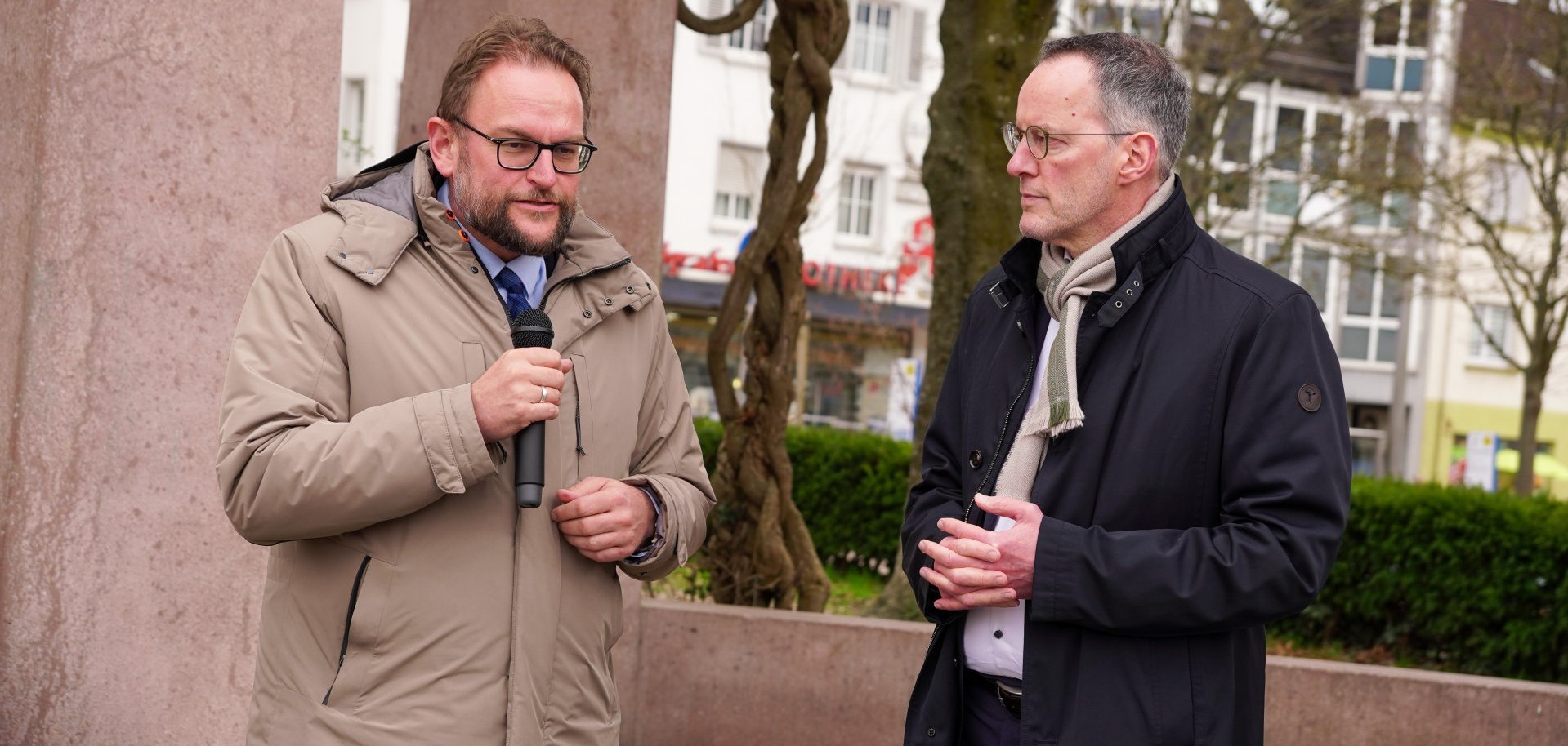 Der Oberbürgermeister von Pirmasens, Markus Zwick, steht links und spricht in ein Handmikrofon. Rechts daneben steht der rheinland-pfälzische Innenminister Michael Ebling, der aufmerksam zuhört. Beide tragen Mäntel und befinden sich im Freien auf einem Platz mit Bäumen und Gebäuden im Hintergrund.