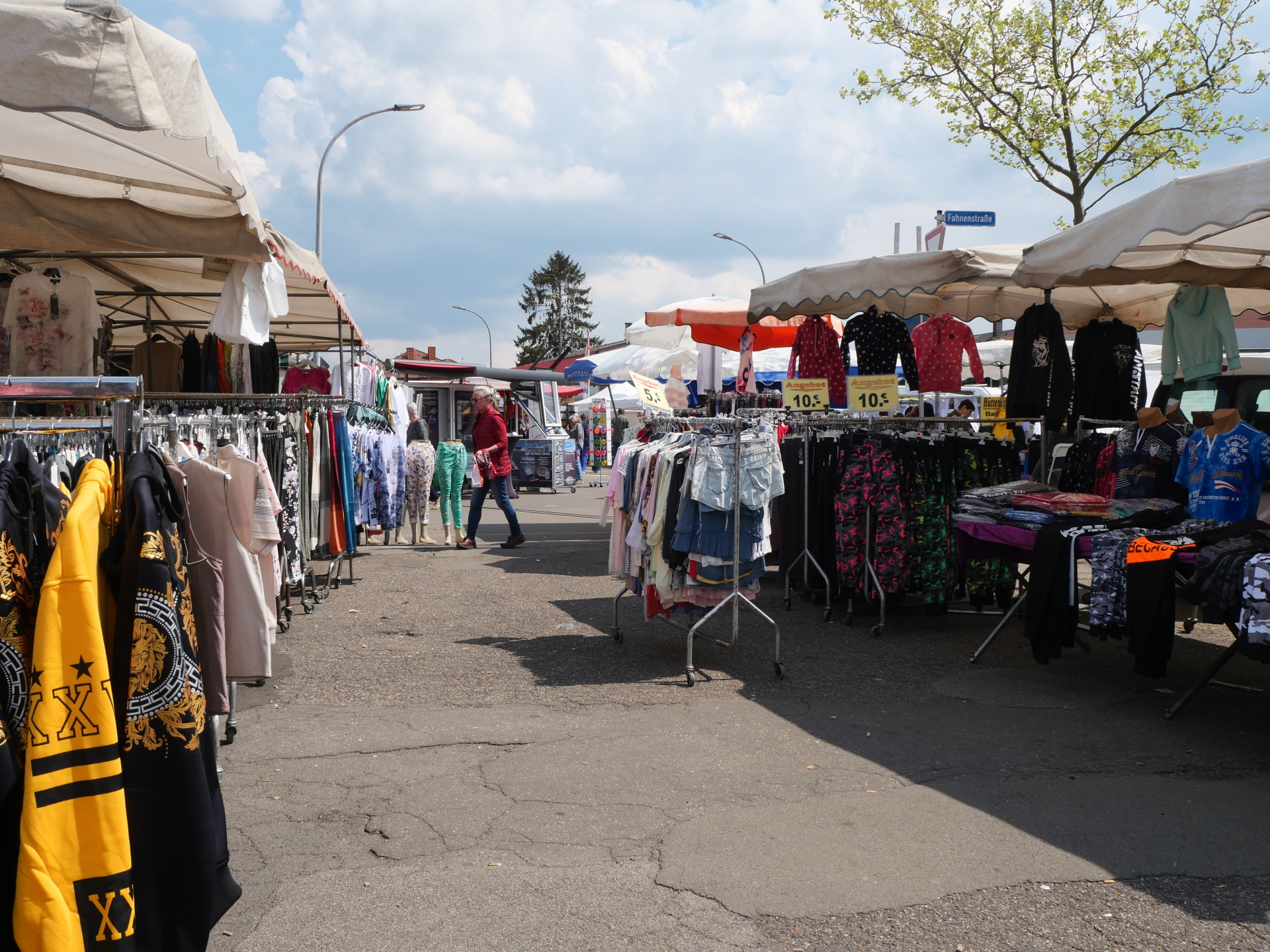 Marktstraße mit mehreren Ständen unter weißen Pavillons, an denen Kleidung auf Kleiderständern hängt; einige Menschen schlendern zwischen den Ständen bei sonnigem Wetter.