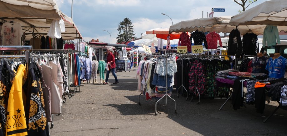 Marktstraße mit mehreren Ständen unter weißen Pavillons, an denen Kleidung auf Kleiderständern hängt; einige Menschen schlendern zwischen den Ständen bei sonnigem Wetter.