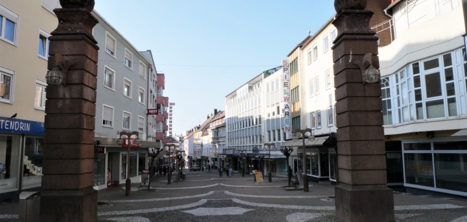 Flammende Granaten südliche Hauptstraße Das Bild zeigt den Eingangsbereich der Fußgängerzone in Pirmasens. Zwei markante Sandsteinpfeiler mit historischen Leuchten rahmen den Blick in die Einkaufsstraße ein. Die Fußgängerzone ist mit einem hellen und dunklen Pflastermuster gestaltet und wird von mehrgeschossigen Geschäfts- und Wohngebäuden gesäumt. Entlang der Straße stehen moderne Straßenlaternen sowie einzelne Passanten, die durch die Innenstadt gehen. Im Hintergrund sind verschiedene Ladenfassaden und Geschäftsschilder zu erkennen. Über der Szene spannt sich ein klarer, wolkenloser Himmel, der der Aufnahme eine helle und freundliche Atmosphäre verleiht.