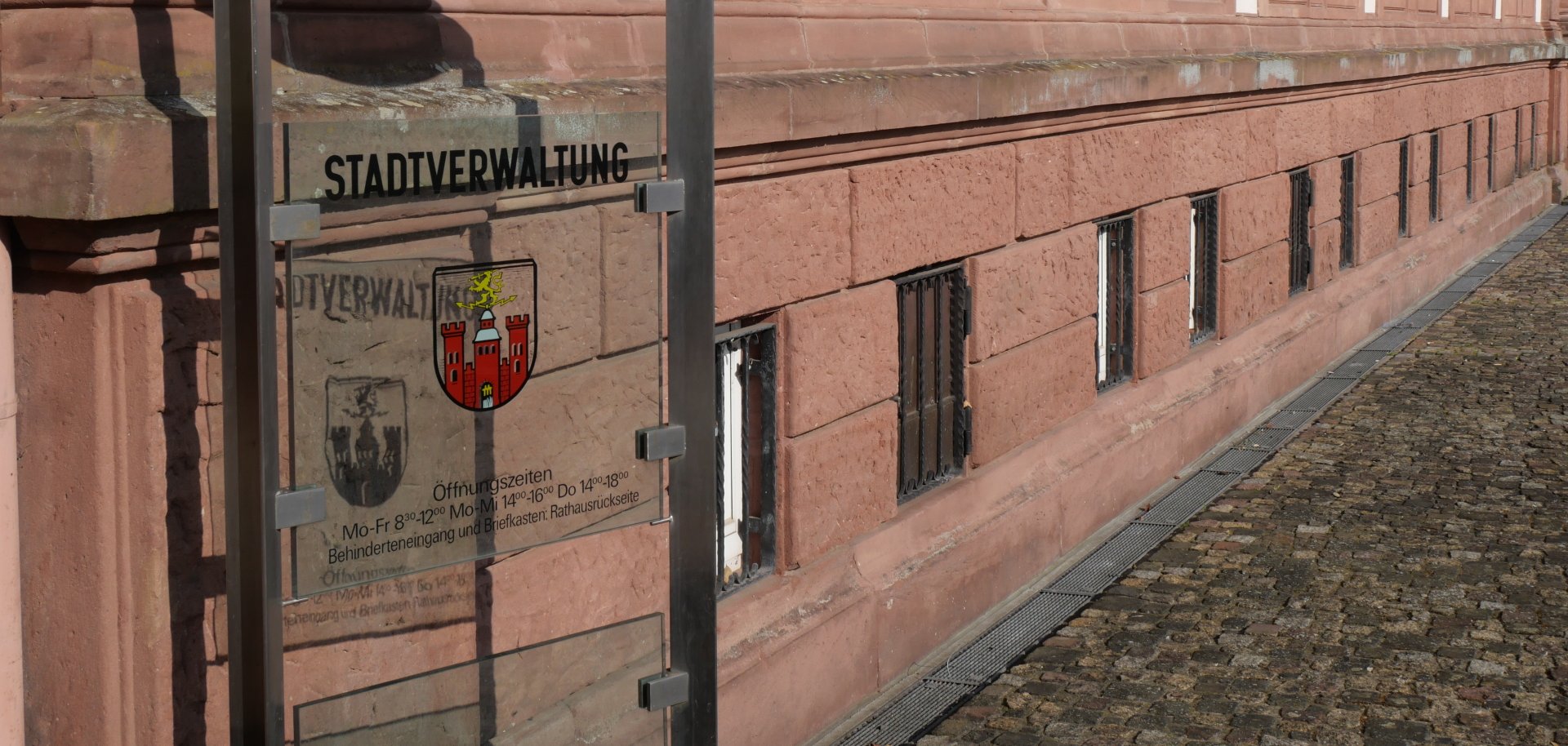 Ein Infoschild der Stadtverwaltung steht vor der historischen Fassade eines Verwaltungsgebäudes mit Blumenkästen an den Fenstern und gepflastertem Gehweg.