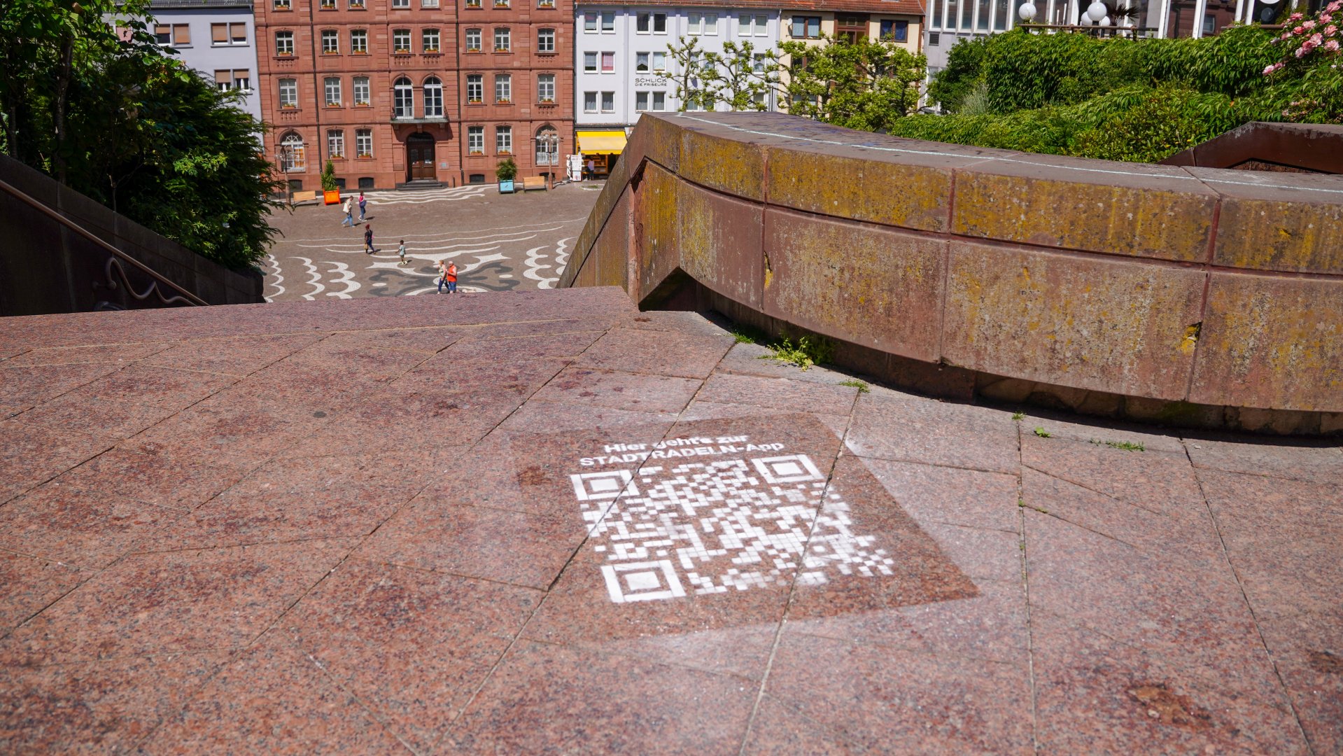 QR-Code Stadtradeln App Blick von einer Treppenanlage auf den Exerzierplatz in Pirmasens. Im Vordergrund ist ein auf den Boden projizierter QR-Code zur „Stadtrallye“-App zu sehen, während im Hintergrund Menschen über den Platz zwischen historischen Gebäuden laufen.