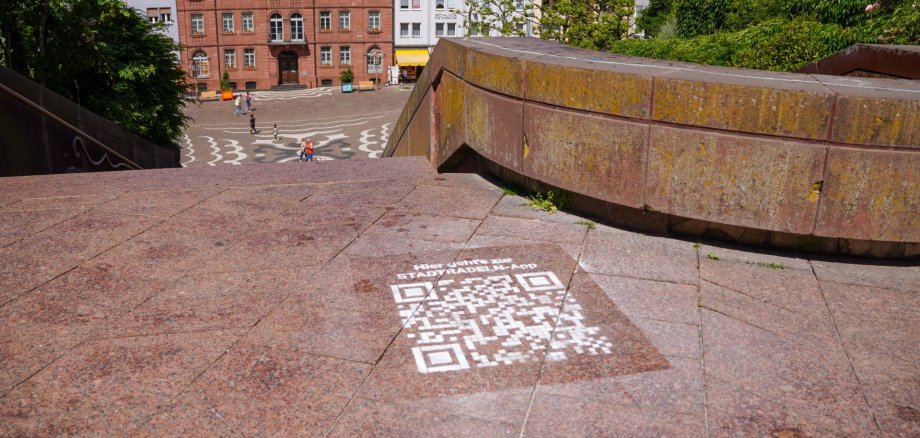 QR-Code Stadtradeln App Blick von einer Treppenanlage auf den Exerzierplatz in Pirmasens. Im Vordergrund ist ein auf den Boden projizierter QR-Code zur „Stadtrallye“-App zu sehen, während im Hintergrund Menschen über den Platz zwischen historischen Gebäuden laufen.