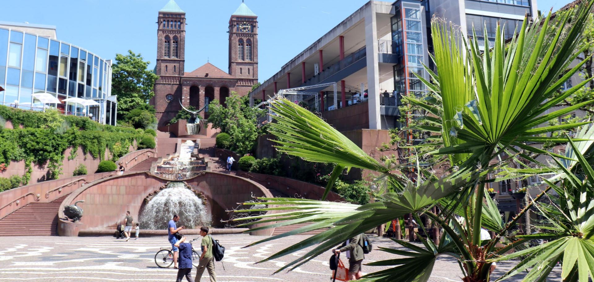 Das Bild zeigt den Schloßbrunnen auf dem Exerzierplatz in Pirmasens an einem sonnigen Tag. Im Vordergrund rahmen die großen grünen Blätter einer Palme die Szene ein. Im Zentrum sprudelt der mehrstufige Brunnen vor der markanten Skulptur, die über eine breite Freitreppe zur Kirche St. Pirmin führt. Die beiden Türme der Kirche ragen vor einem strahlend blauen Himmel empor. Rechts und links säumen moderne Gebäude den Platz, während mehrere Passanten die Fußgängerzone überqueren oder sich am Brunnen aufhalten. Die Aufnahme vermittelt eine sommerliche Atmosphäre und verbindet historische Architektur mit dem lebendigen Stadtzentrum.