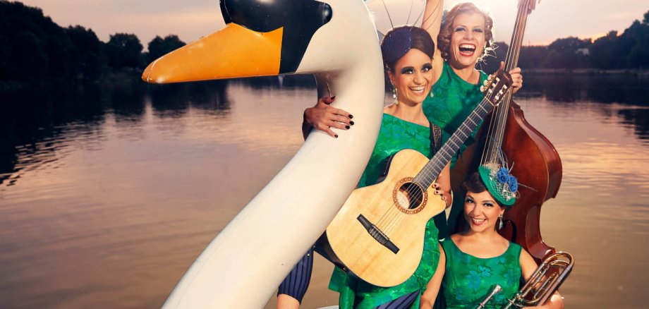 Drei Musikerinnen in leuchtend grünen Retro-Kleidern posieren auf einem großen weißen Schwanen-Tretboot auf einem See. Eine Frau hält eine Gitarre, eine weitere steht mit einem Kontrabass hinter ihr und hebt lachend einen Arm in die Luft, während die dritte mit einer Posaune im vorderen Bereich des Bootes sitzt. Die Frauen tragen auffällige Kopfschmuck-Accessoires und präsentieren sich gut gelaunt im Stil einer nostalgischen Bühnenshow. Im Hintergrund ist die untergehende Sonne über dem ruhigen See zu sehen.